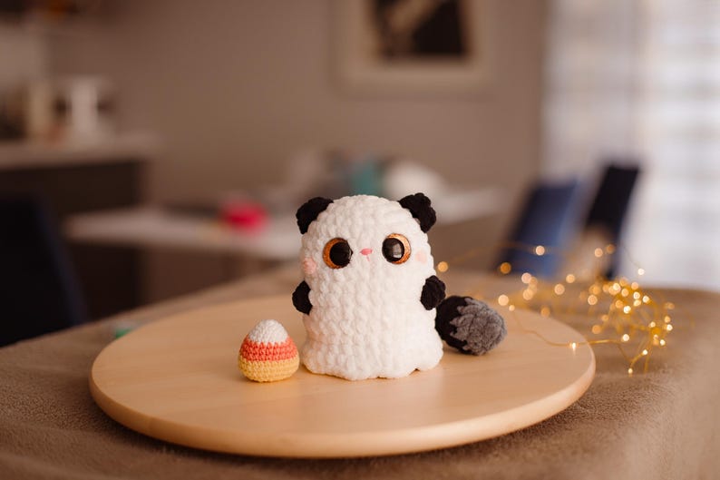 May include: A white crocheted ghost with black ears and a tail, holding a candy corn. The ghost has large, black eyes and a pink nose. The ghost and candy corn are sitting on a wooden surface.