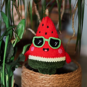 Puede incluir: Un juguete de peluche de sand&iacute;a de color rojo y verde con gafas de sol. La sand&iacute;a tiene una cara sonriente y est&aacute; sentada en una cesta tejida.