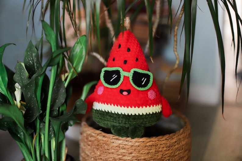 Puede incluir: Un juguete de peluche de sand&iacute;a de color rojo y verde con gafas de sol. La sand&iacute;a tiene una cara sonriente y est&aacute; sentada en una cesta tejida.