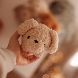 May include: A soft, cream-colored plush toy shaped like a dog with black eyes. The toy is being held in a hand.