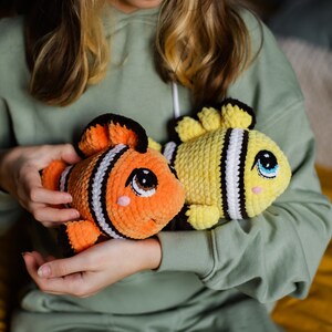 May include: Two crocheted fish toys, one orange and black with white stripes and one yellow with black stripes, are held by a person.