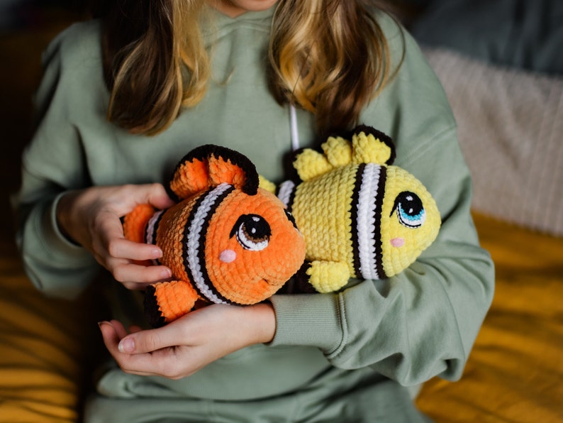 May include: Two crocheted fish toys, one orange and black with white stripes and one yellow with black stripes, are held by a person.