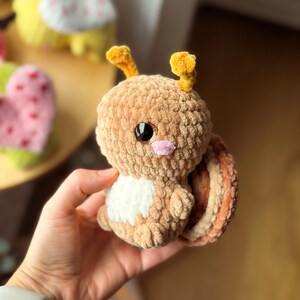 May include: A handmade, plush snail toy in shades of brown, tan, and white. The snail has large black eyes, a pink nose, and yellow antennae. The shell is striped with varying shades of brown and tan. The toy is soft and cuddly.