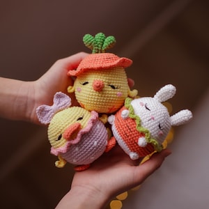 May include: Three crocheted stuffed animals: a yellow chick with a green stem and orange carrot, a yellow chick with a purple and white shell, and a white bunny with an orange carrot and green leaves.