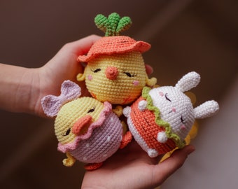 May include: Three crocheted stuffed animals: a yellow chick with a green stem and orange carrot, a yellow chick with a purple and white shell, and a white bunny with an orange carrot and green leaves.