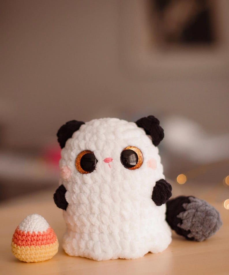 May include: White crocheted ghost plush toy with black ears and tail, holding a crocheted candy corn.