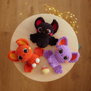 May include: Three crocheted bat toys in orange, purple and black. The bats have large eyes and are sitting on a wooden surface.