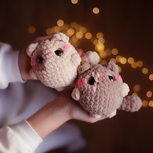 May include: Two crocheted cat toys, one beige and one brown, with pink cheeks and black eyes. The toys are being held by a person's hands.