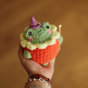 May include: A green crocheted frog wearing a purple and white hat and holding a gold wand. The frog is sitting on a yellow and orange crocheted base.