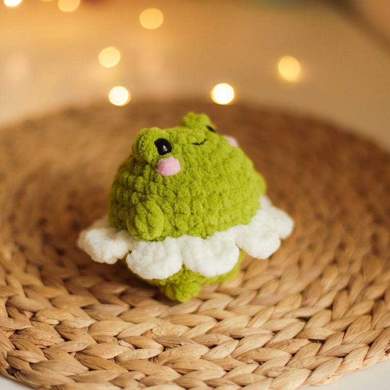 May include: A green crocheted frog wearing a white crocheted tutu. The frog has pink cheeks and is sitting on a woven wicker surface.