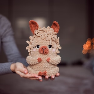 May include: A handmade, plush llama toy in shades of tan and brown. The llama has a fluffy mane, small ears, and a sweet expression with rosy cheeks. The toy is held in a person's hands, showcasing its soft texture and detailed craftsmanship.