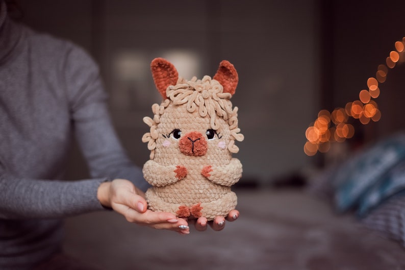 May include: A handmade, plush llama toy in shades of tan and brown. The llama has a fluffy mane, small ears, and a sweet expression with rosy cheeks. The toy is held in a person's hands, showcasing its soft texture and detailed craftsmanship.