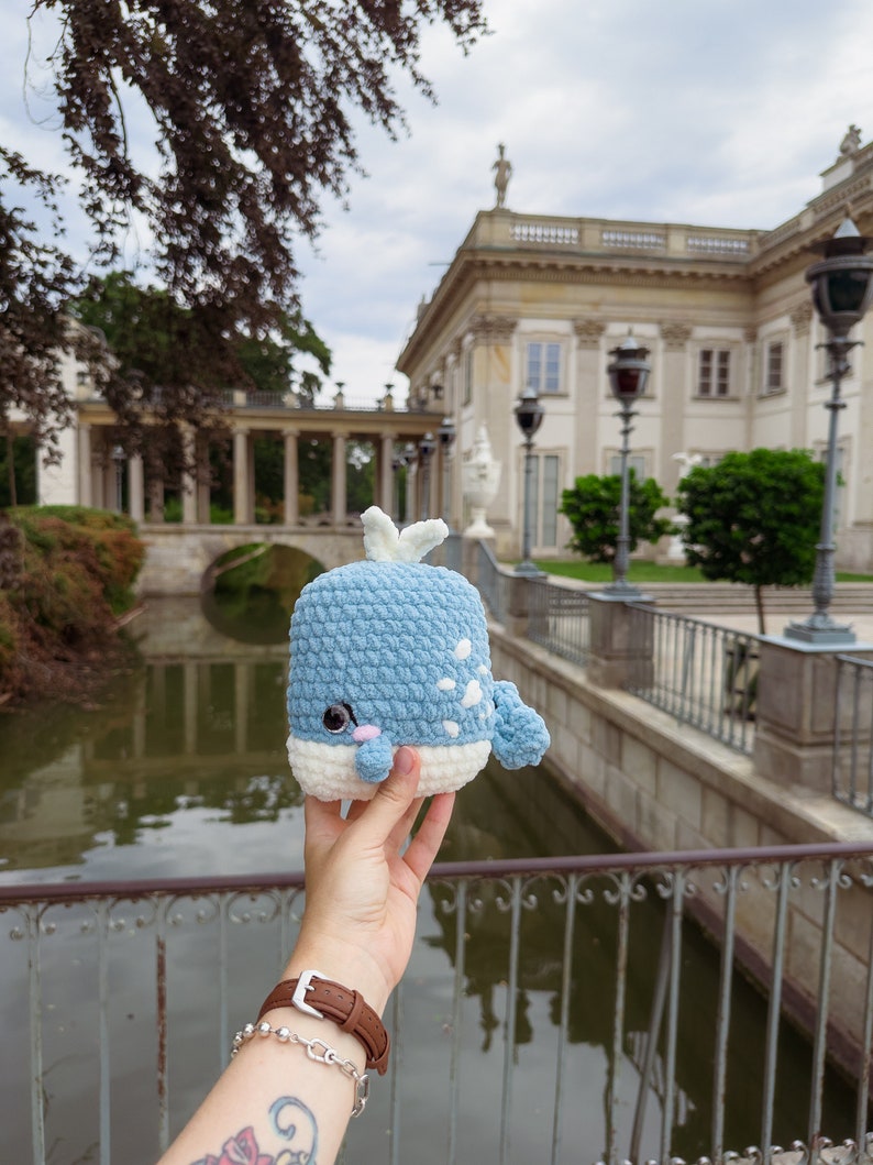 May include: A blue and white crocheted whale plush toy held in front of a classical building with a pond in the foreground.