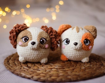 May include: Two handmade crochet dog toys. One dog has brown and white fur with curly brown ears. The other dog has white, orange, and brown fur. Both have large, expressive eyes and pink cheeks. They sit on a woven mat.