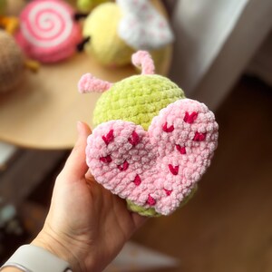 May include: A handmade, crocheted toy shaped like a green bug with a pink heart. The heart is covered in small, red heart-shaped accents. Other crocheted toys are visible in the background, including a pink and white swirl and a yellow bug.