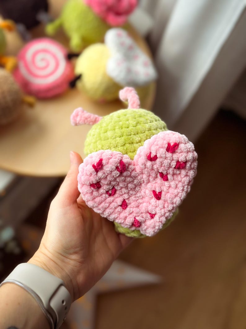 May include: A handmade, crocheted toy shaped like a green bug with a pink heart. The heart is covered in small, red heart-shaped accents. Other crocheted toys are visible in the background, including a pink and white swirl and a yellow bug.