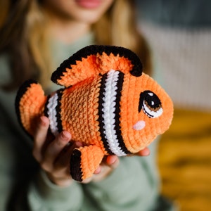 May include: A crocheted orange and black clownfish toy with white stripes and a cute face. The toy is being held by a person.