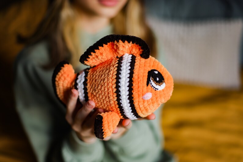 May include: A crocheted orange and black clownfish toy with white stripes and a cute face. The toy is being held by a person.