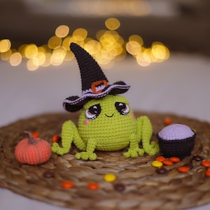 Pode incluir: Um sapo de crochê verde usando um chapéu de bruxa preto com uma faixa roxa e uma pequena abóbora laranja. O sapo está sentado em um tapete de vime marrom com doces roxos e marrons.