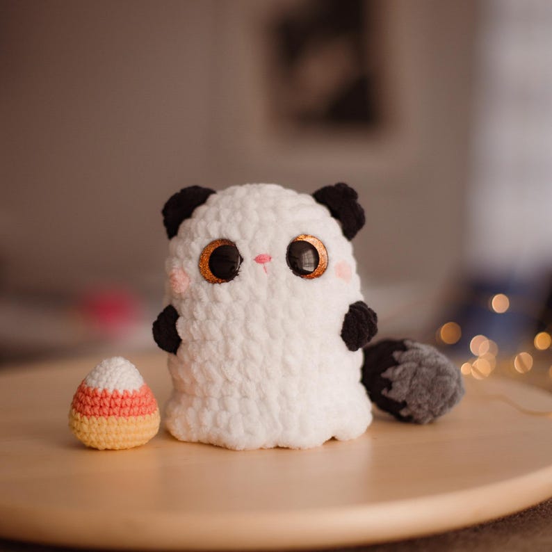 May include: White crocheted ghost plush toy with black ears and a gray tail. The ghost has large black eyes with gold pupils and a pink nose. The ghost is sitting on a wooden surface next to a candy corn.