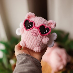 May include: A pink crocheted pig wearing heart-shaped sunglasses. The pig has white wings and is being held by a hand.