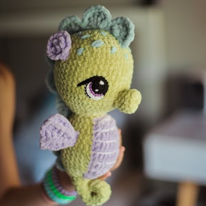 May include: A crocheted seahorse toy with a green body, purple fins, and blue and green details on its head. The seahorse has large, black eyes with purple eyelids.