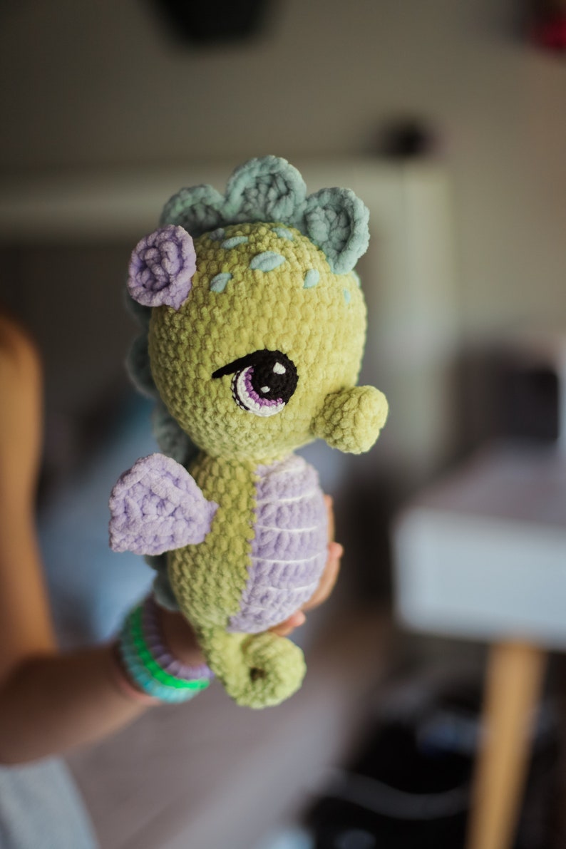 May include: A crocheted seahorse toy with a green body, purple fins, and blue and green details on its head. The seahorse has large, black eyes with purple eyelids.