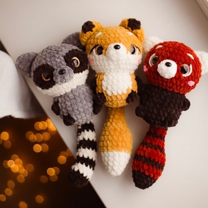 May include: Three handmade crochet animal toys: a gray raccoon with a striped tail, a yellow and white fox, and a red panda with a striped tail. Each toy has large, expressive eyes and is made of soft, textured yarn.