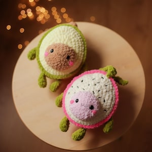 May include: Two crocheted stuffed toys shaped like fruit. One is a green avocado with a brown pit and the other is a pink dragon fruit with white flesh. Both have smiling faces.
