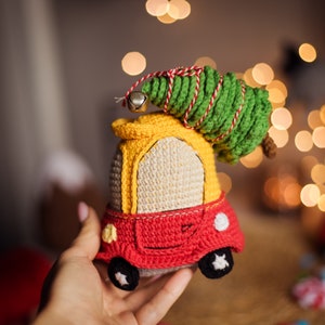 Può includere: Un'auto giocattolo all'uncinetto in rosso, giallo e bianco con un albero di Natale all'uncinetto verde in cima. L'auto ha un campanello sul davanti.
