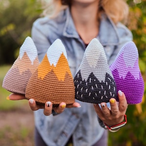 Op de afbeelding: Vier gehaakte pluche speelgoed in de vorm van bergen in verschillende kleuren. De bergen zijn bruin, oranje, grijs en paars. Het speelgoed wordt in de handen van een persoon gehouden.