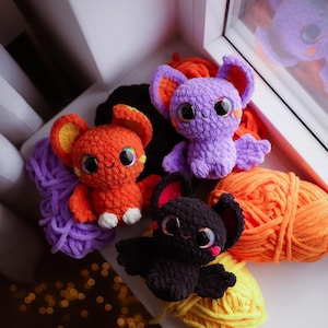Puede incluir: Cuatro peluches de murci&eacute;lago de crochet en diferentes colores: morado, naranja, negro y una bola de lana morada. Los peluches tienen ojos grandes y est&aacute;n sentados sobre una superficie blanca con bolas de lana naranja y amarilla.