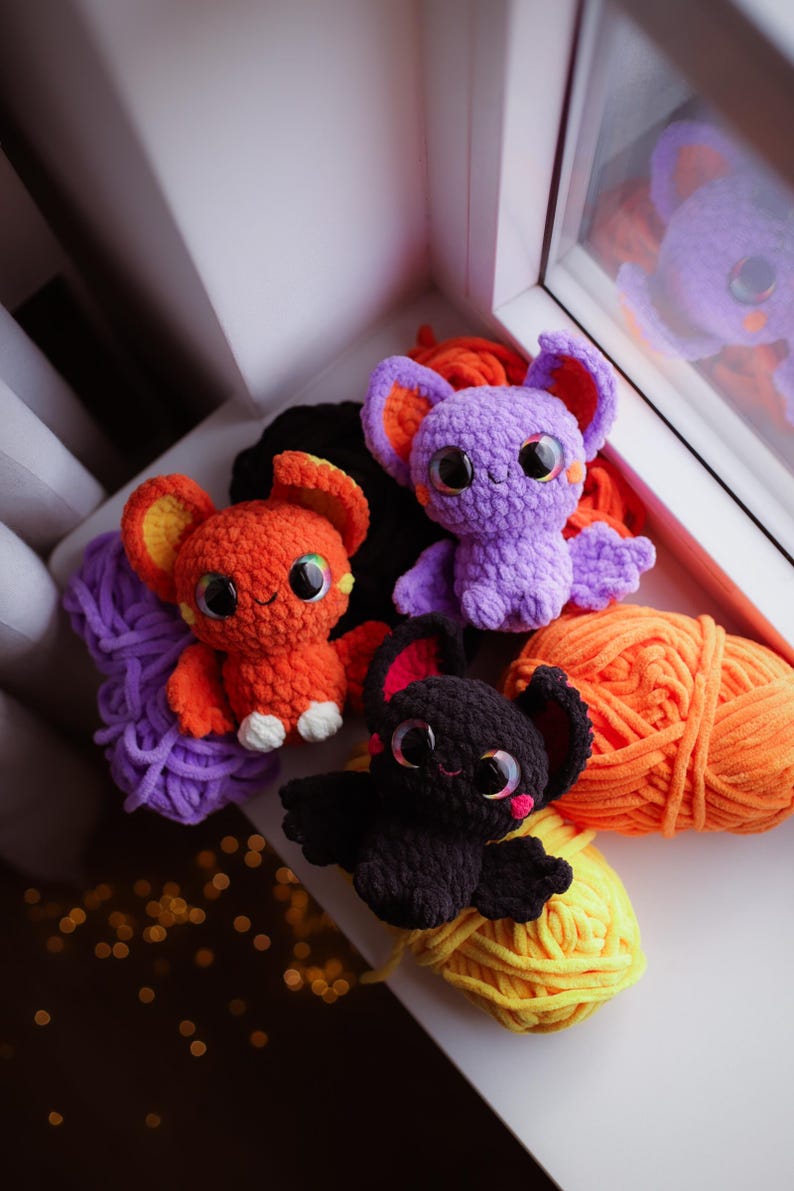 Puede incluir: Cuatro peluches de murci&eacute;lago de crochet en diferentes colores: morado, naranja, negro y una bola de lana morada. Los peluches tienen ojos grandes y est&aacute;n sentados sobre una superficie blanca con bolas de lana naranja y amarilla.