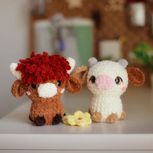 Op de afbeelding: Twee gehaakte knuffels, een bruine koe met rood krullend haar en een witte koe met bruine oren en een roze neus. De koeien zitten op een wit oppervlak met een gele bloem tussen hen in.