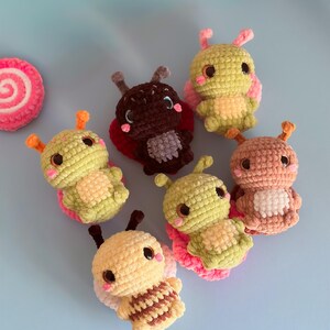 May include: A collection of handmade crochet insect toys in various colors, including yellow, green, brown, and peach. Each toy features large eyes, antennae, and wings. A pink and white spiral-shaped crochet item is also present.