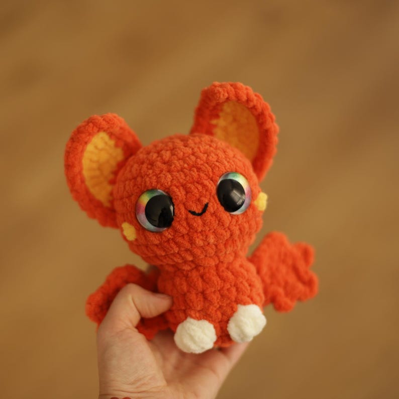 Puede incluir: Un murci&eacute;lago naranja de crochet con orejas amarillas y pies blancos. El murci&eacute;lago tiene ojos grandes y coloridos y una expresi&oacute;n sonriente.
