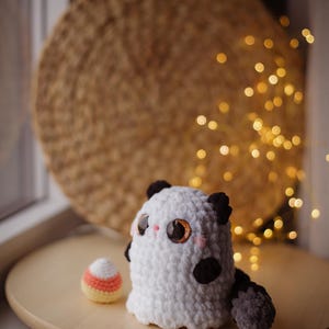 May include: A white crocheted ghost with black eyes and a black and gray tail sits on a wooden surface next to a crocheted candy corn.