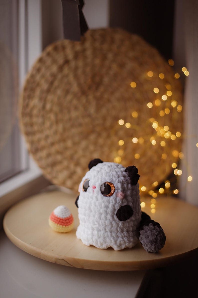 May include: A white crocheted ghost with black eyes and a black and gray tail sits on a wooden surface next to a crocheted candy corn.
