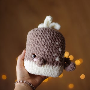 May include: A crocheted whale plush toy with brown and white yarn. The whale has a white belly and a brown back with a white spout on top. The whale has a cute face with black eyes and a pink nose.