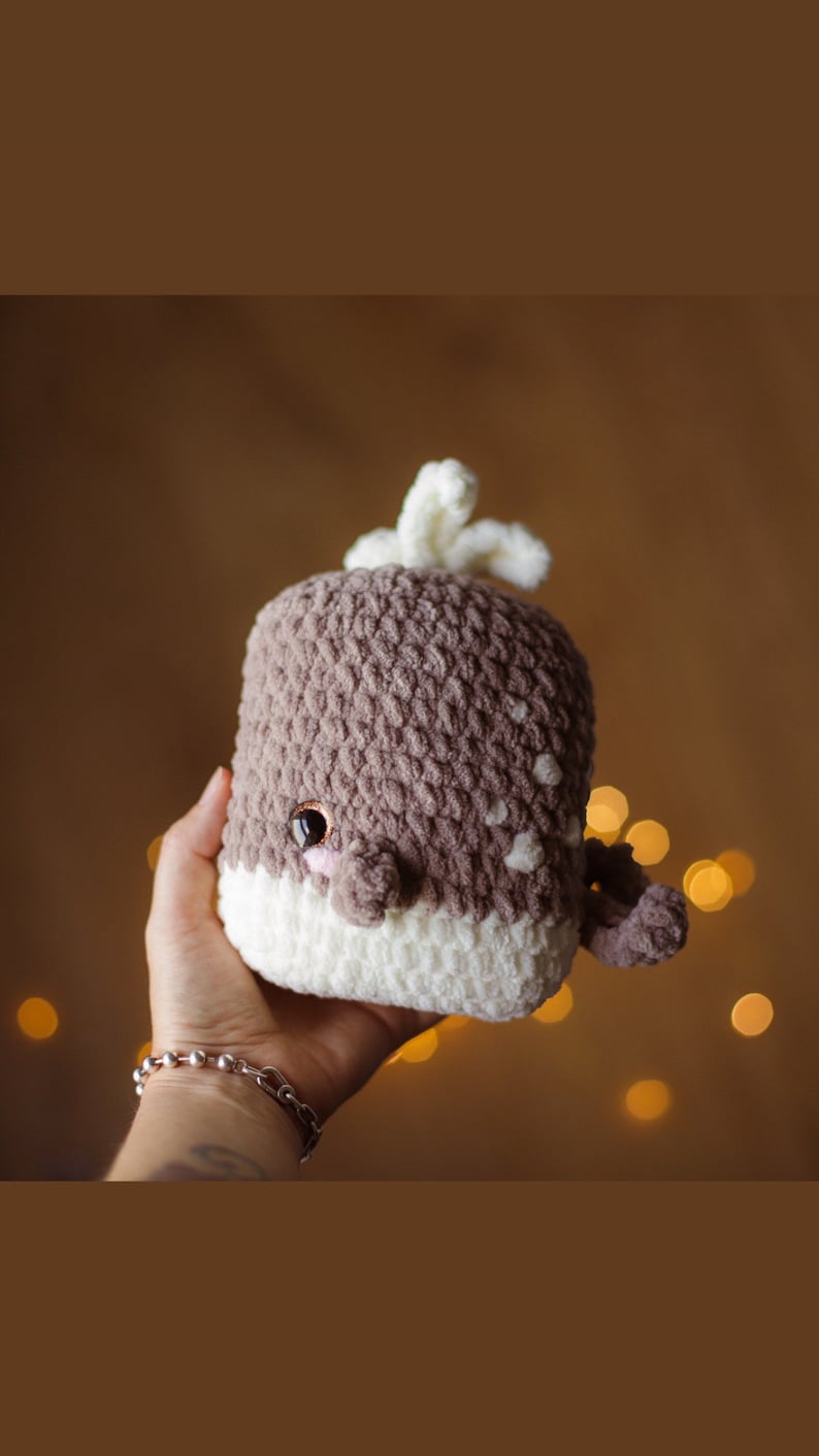 May include: A crocheted whale plush toy with brown and white yarn. The whale has a white belly and a brown back with a white spout on top. The whale has a cute face with black eyes and a pink nose.