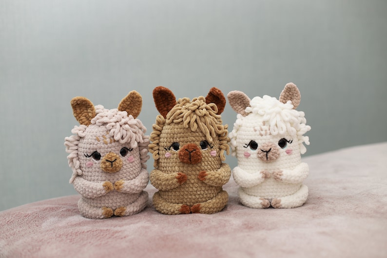 May include: Three handmade crochet alpaca dolls in soft pastel colors. The alpaca dolls are in shades of pink, tan, and white, each with detailed features like ears, eyes, and a fluffy mane. They are sitting upright with their hands clasped.