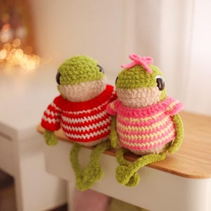 May include: Two handmade crochet frog dolls. One frog wears a red and white striped sweater, the other wears a pink and yellow striped sweater and a pink bow. Both frogs have green heads and legs, and black eyes.