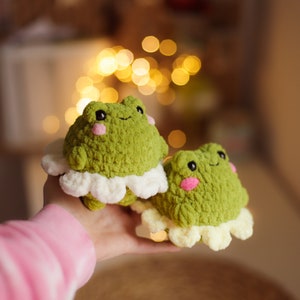 May include: Two green crocheted frog plushies with white crocheted ruffles around their bodies. The frogs have pink cheeks and black eyes.