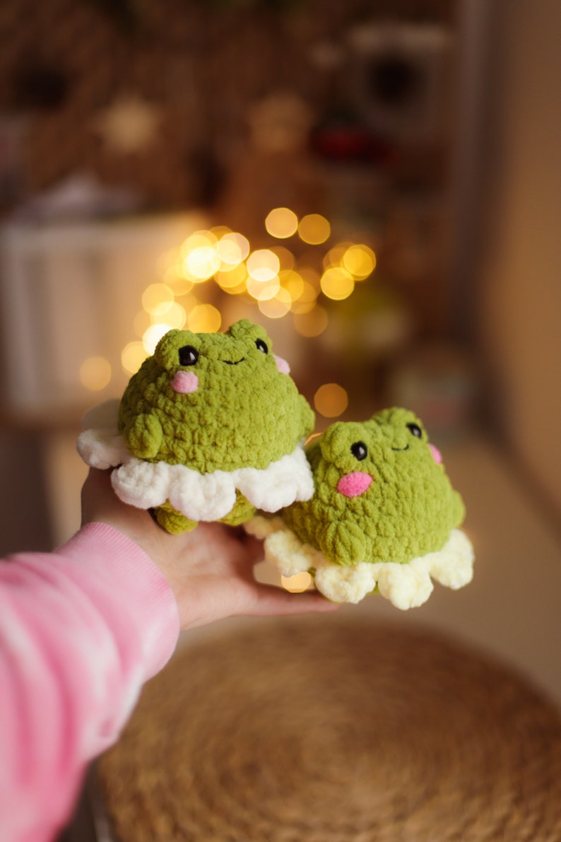 May include: Two green crocheted frog plushies with white crocheted ruffles around their bodies. The frogs have pink cheeks and black eyes.