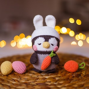 Pode incluir: Um pinguim de crochê usando um chapéu de coelho branco e segurando uma cenoura. O pinguim está sentado em uma cesta de vime com dois ovos de Páscoa rosa e amarelos.