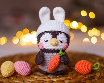 May include: A crocheted penguin wearing a white bunny hat and holding a carrot. The penguin is sitting on a woven basket with two pink and yellow Easter eggs.