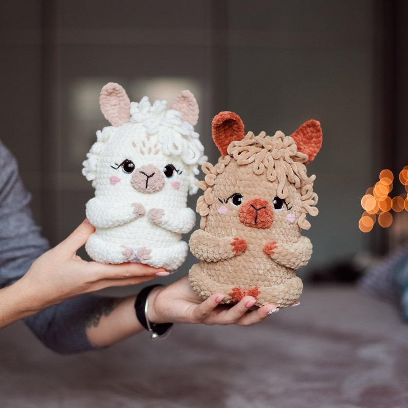 May include: Two handmade crochet llama dolls. One is white with pink accents, the other is tan with brown accents. Both have detailed facial features, including eyes, noses, and small arms. The dolls are held in a person's hands.