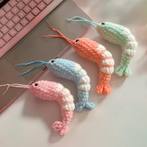 Könnte beinhalten: Vier gehäkelte Garnelen-Spielzeuge in Pastellfarben: Rosa, Blau, Orange und Grün. Jede Garnele hat weiße Streifen und blaue oder orangefarbene Antennen. Die Spielzeuge sind auf einer hellen Oberfläche in der Nähe eines Laptops angeordnet.