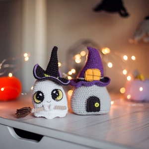 May include: Two crocheted Halloween decorations, a ghost wearing a witch's hat and a house with a purple pointed roof. The ghost is holding a broom and has big eyes. The house has a small door and a window. Both decorations are on a white surface with string lights.