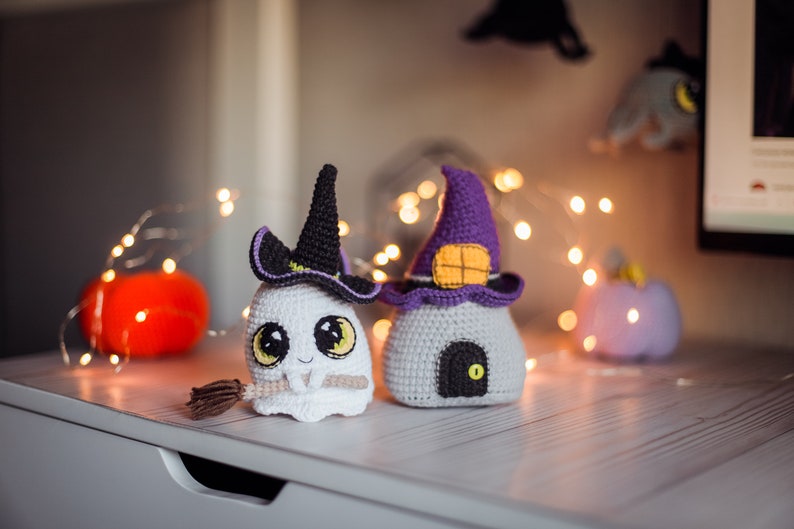 May include: Two crocheted Halloween decorations, a ghost wearing a witch's hat and a house with a purple pointed roof. The ghost is holding a broom and has big eyes. The house has a small door and a window. Both decorations are on a white surface with string lights.